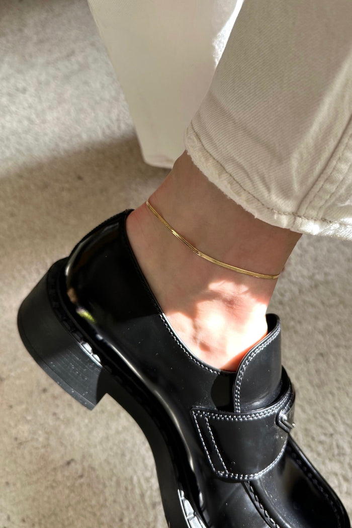 A gold-colored mini herringbone anklet on a person's ankle, visible above beige pants and a black shoe.