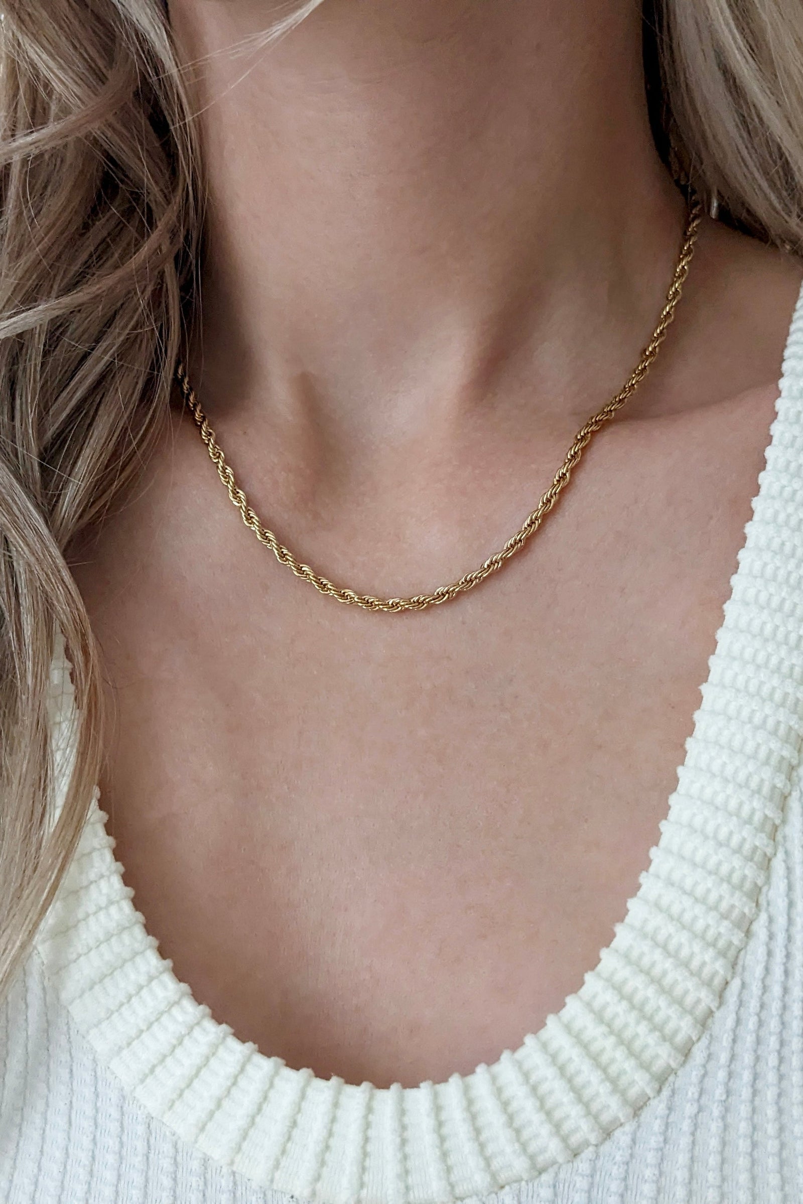 A close-up of a woman wearing a delicate gold chain necklace with a twisted rope design, paired with a white ribbed knit top.