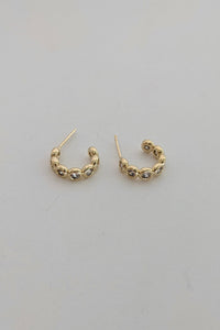 A pair of small gold sparkle hoop earrings with textured, beaded detailing along the front, displayed on a plain light gray surface.