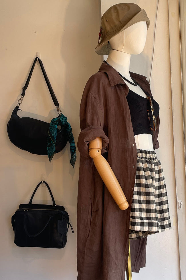 A mannequin dressed in a brown long-sleeve shirt, black crop top, black-and-white gingham skirt, and tan hat, displayed indoors next to two black handbags hanging on a wall.