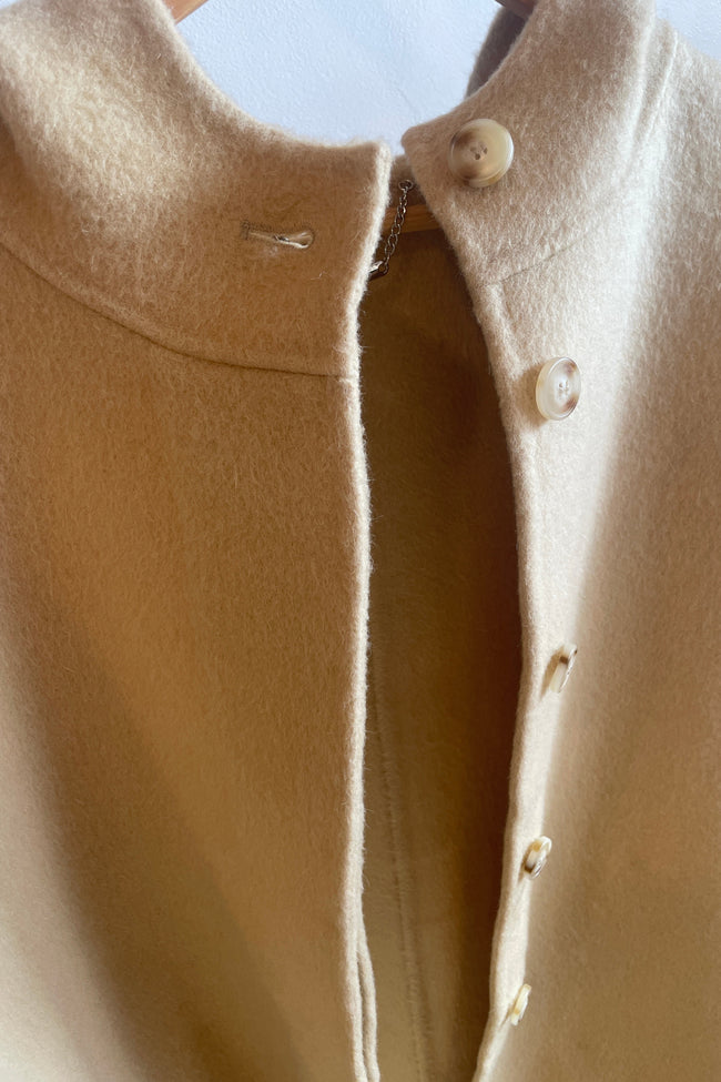 Close-up of a beige wool coat with a high folded collar and decorative buttons down the front, shown hanging against a light background.
