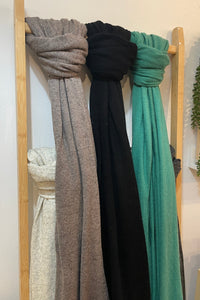 Three knit scarves in taupe, black, and teal green draped over a light wood hanger rack against a plain indoor wall.