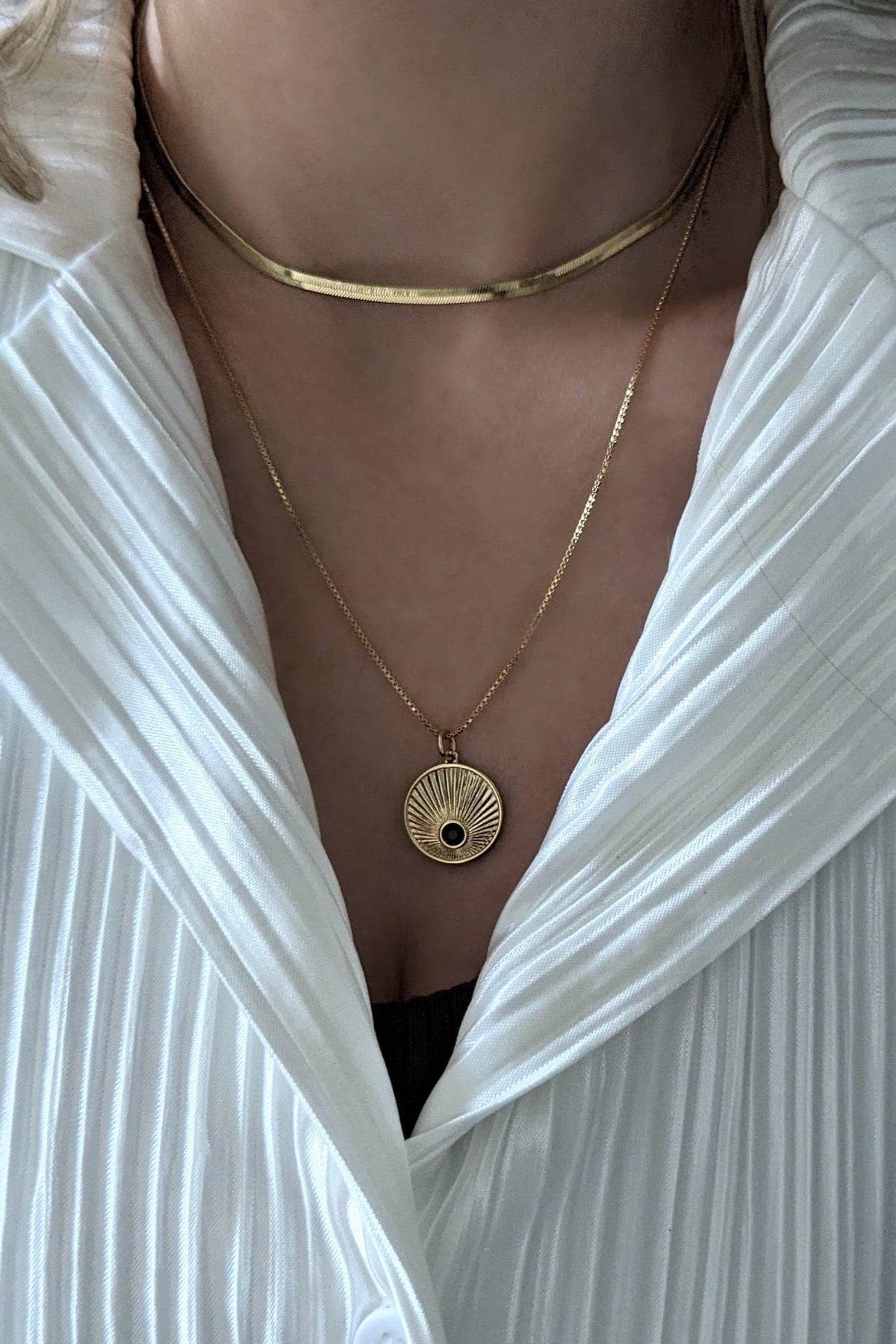 A person wearing a white shirt layered with a gold-filled chain necklace with a starburst pendant and a black cubic zirconia stone.