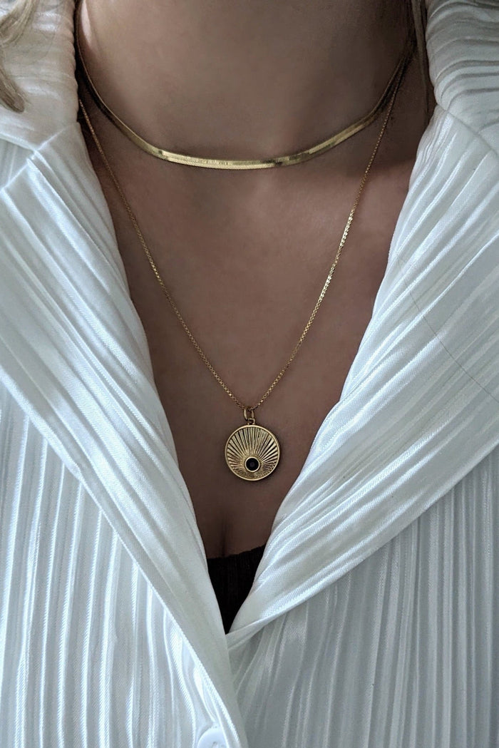 A person wearing a white shirt layered with a gold-filled chain necklace with a starburst pendant and a black cubic zirconia stone.