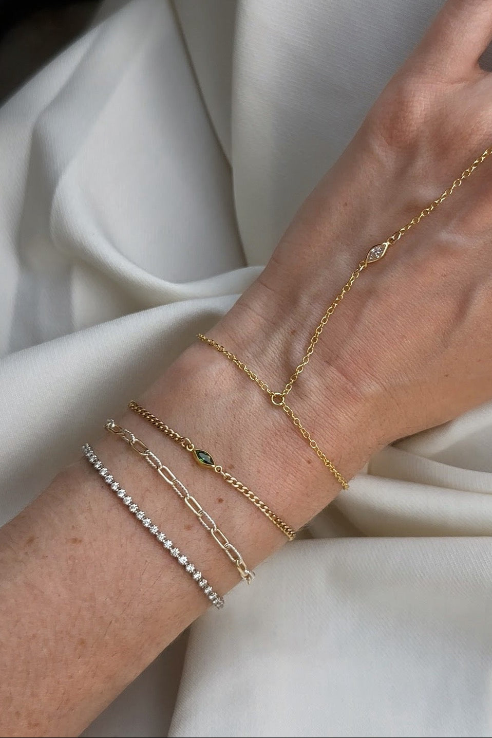 A close-up of a person's wrist and hand wearing multiple delicate gold bracelets and a matching gold chain handpiece, styled against draped white fabric.