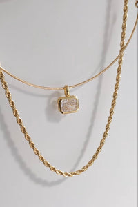 A gold layered necklace set featuring a thin bangle-style top layer with a square bezel-set clear crystal pendant, and a thicker twisted rope chain layer below, displayed on a light gray background.