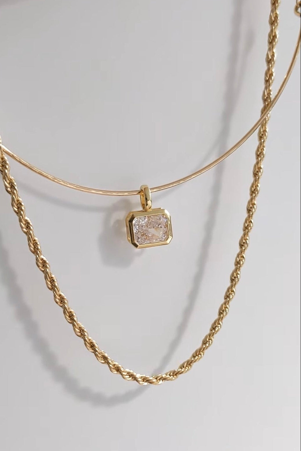 A gold layered necklace set featuring a thin bangle-style top layer with a square bezel-set clear crystal pendant, and a thicker twisted rope chain layer below, displayed on a light gray background.