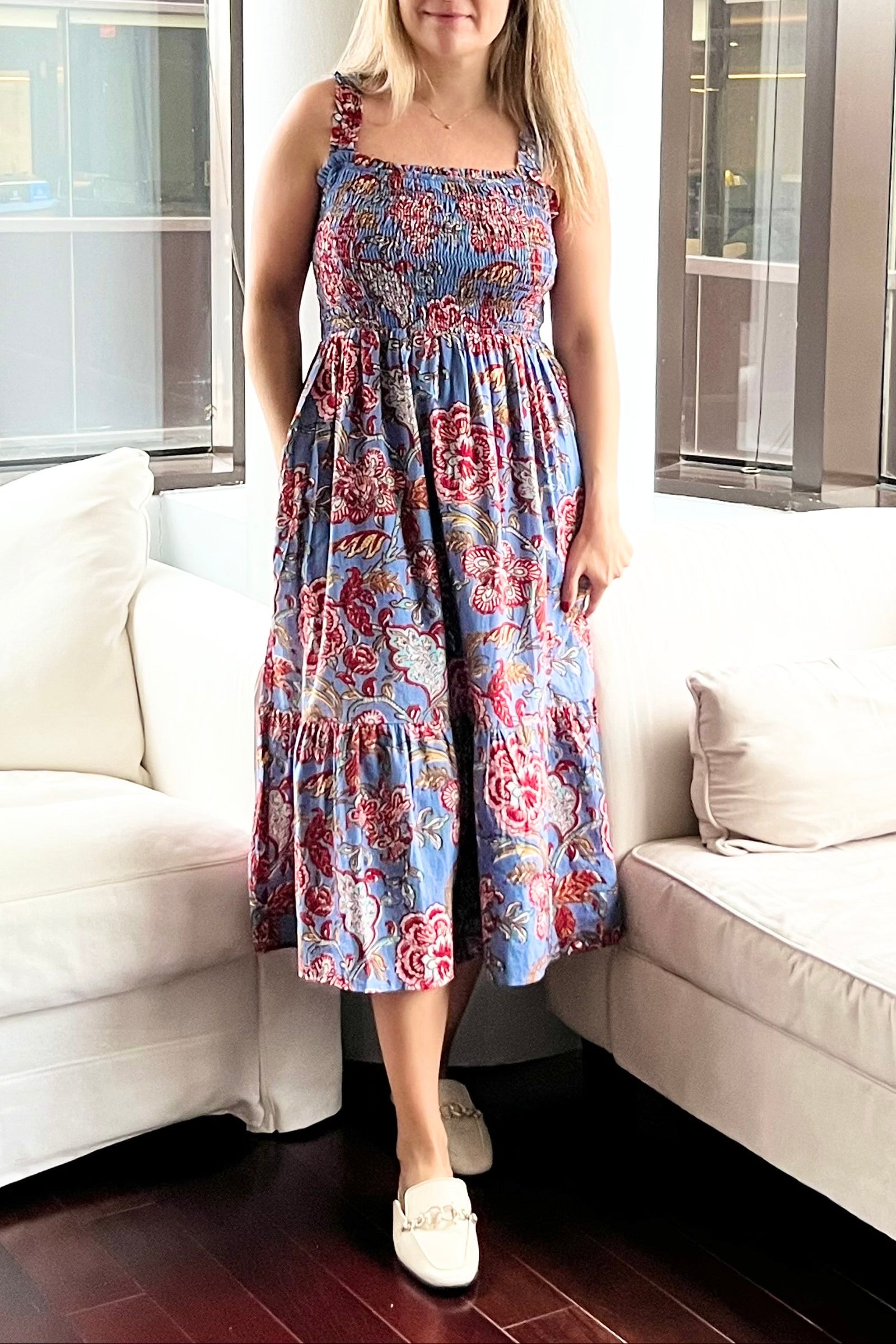 Woman in a floral dress standing in a living room with white couches and large windows.