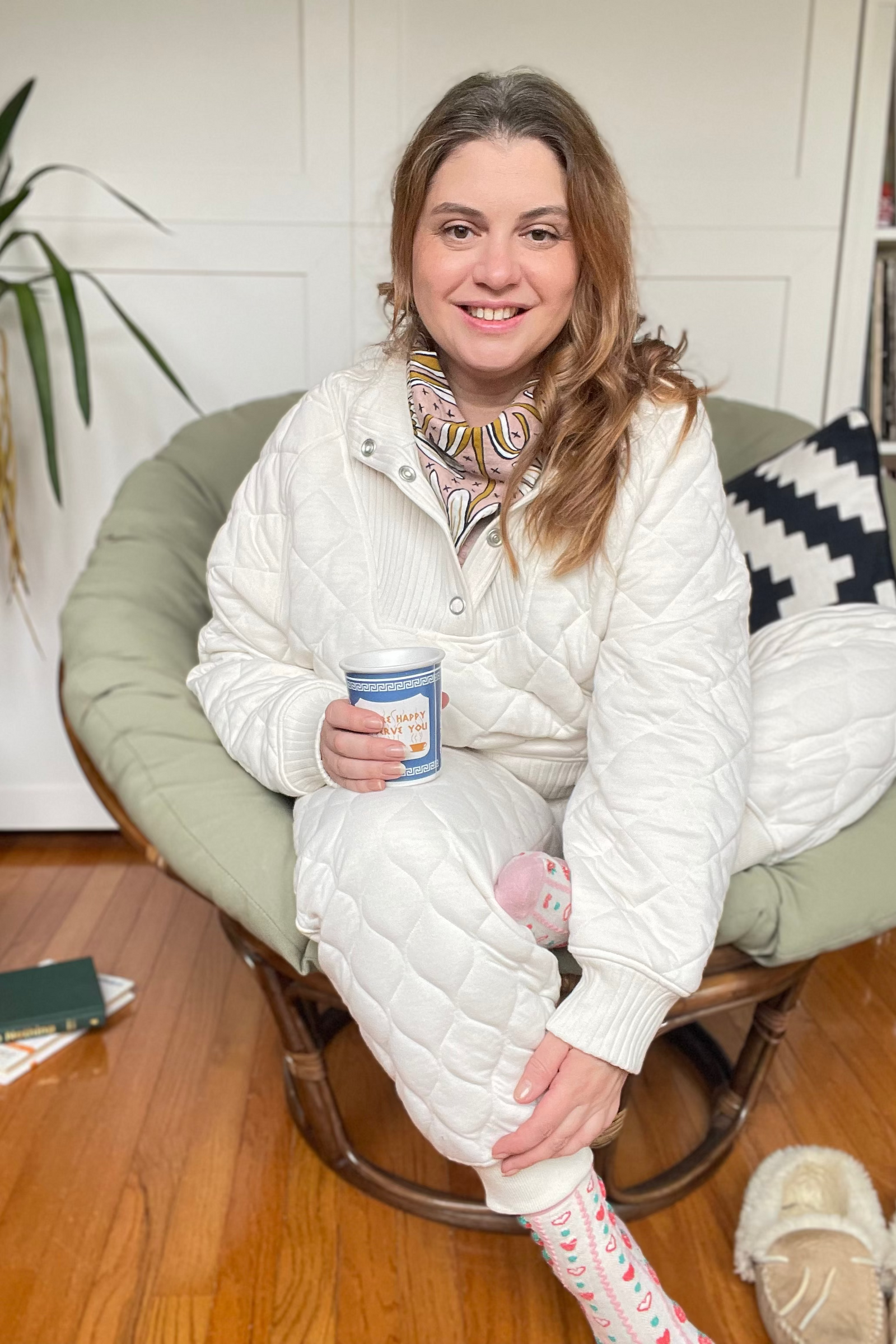 Woman in cloud dancer white loungewear sitting on a green chair holding a coffee cup.