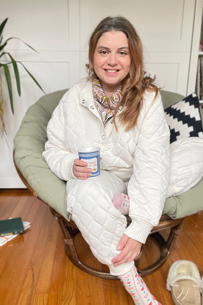 Woman in cloud dancer white loungewear sitting on a green chair holding a coffee cup.
