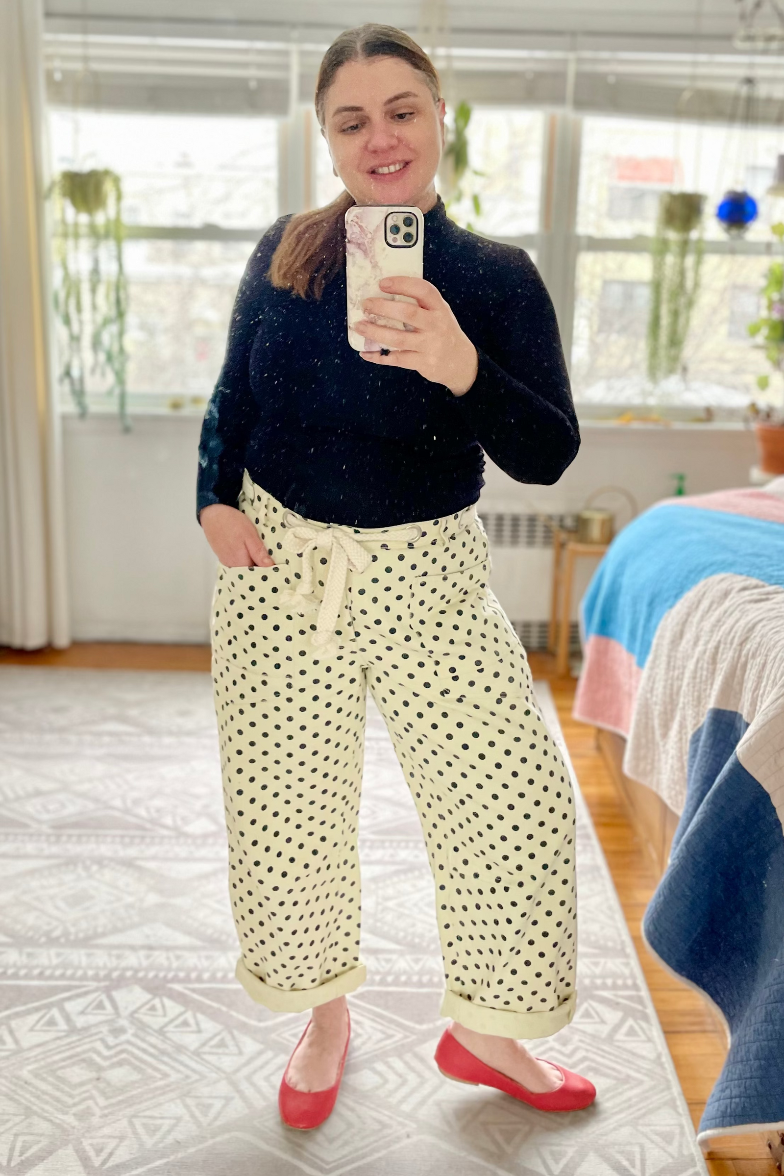 Person taking a mirror selfie wearing a black sweater and cloud dancer white polka dot pants in a bedroom.