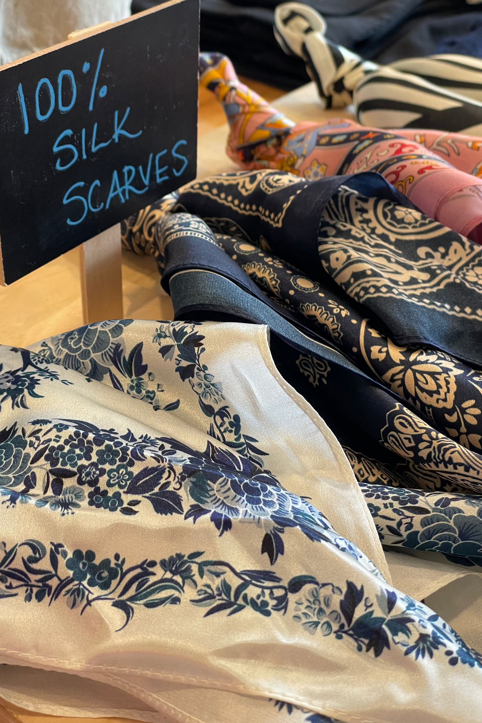Silk scarves with floral and paisley patterns displayed on a table, accompanied by a sign indicating 100% silk.