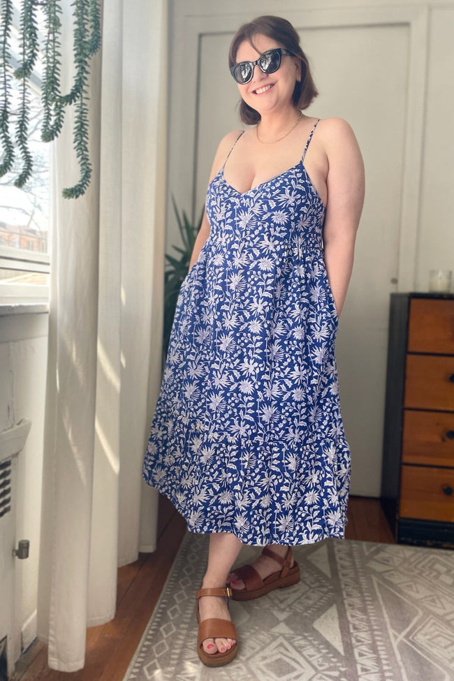 A woman standing in an indoor setting wearing a midi dress with a floral pattern, sleeveless design, and side pockets. She is also wearing brown sandals.