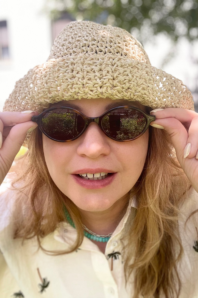 Woman wearing a beige woven hat and sunglasses with a blurred outdoor background