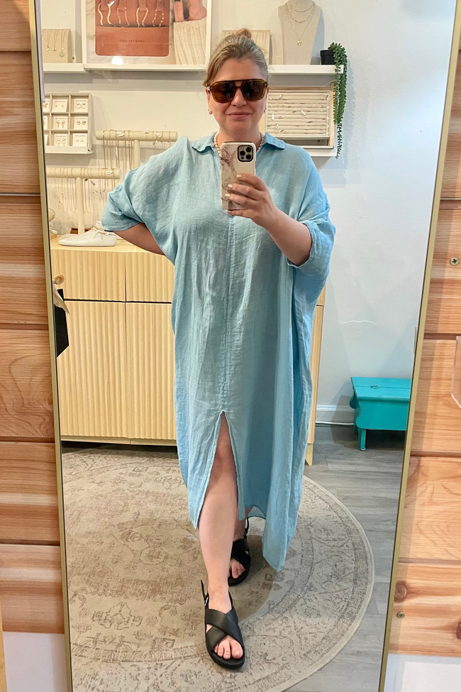 Person wearing a light blue dress taking a mirror selfie in a room with wooden shelves and a rug.