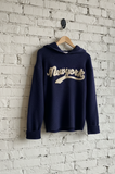 Navy blue hoodie with 'New York' text on a wooden hanger against a white brick wall.