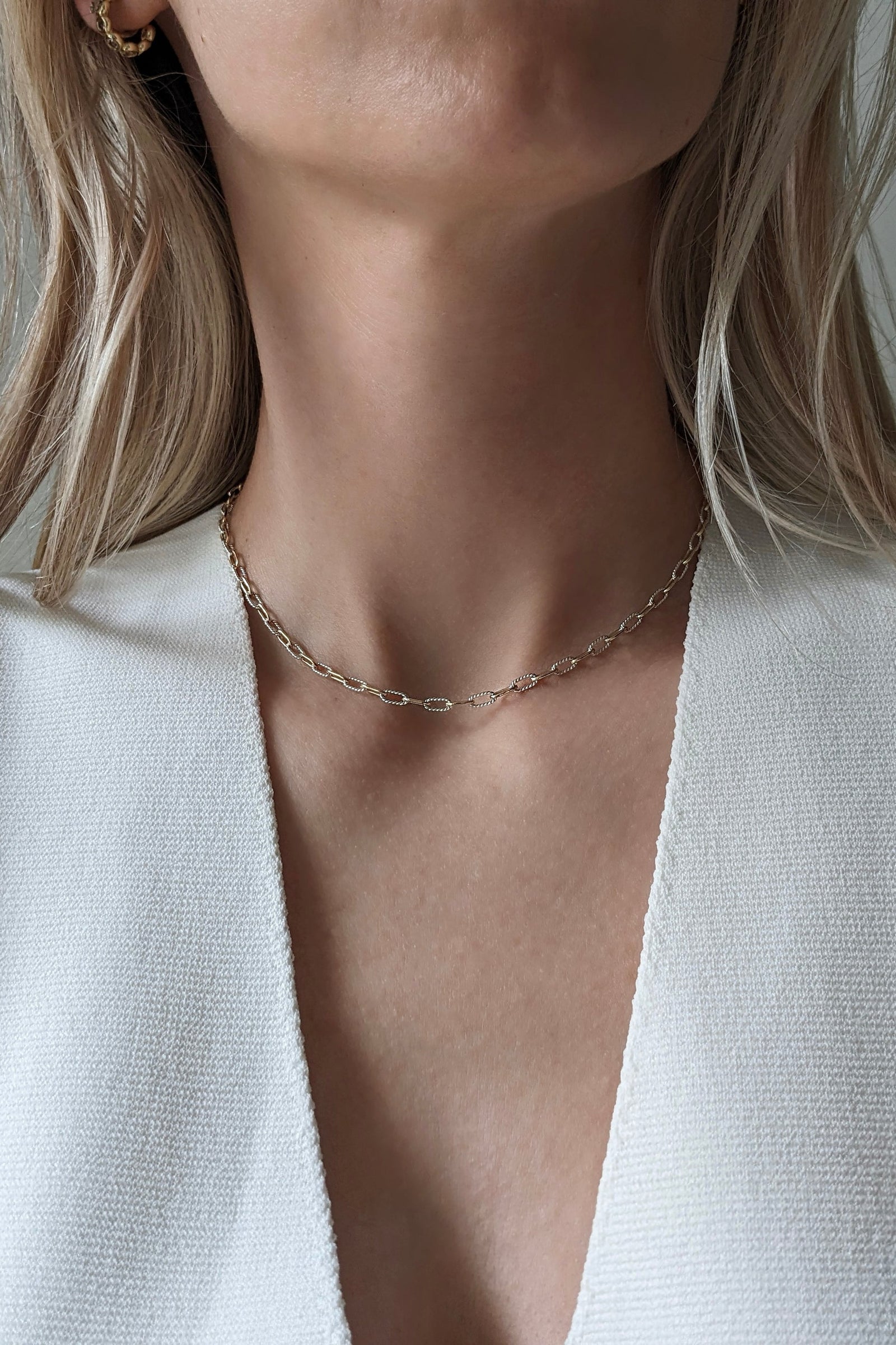 A close-up of a model's neck and upper chest wearing a delicate gold chain necklace with a small paperclip-style link design, paired with a white textured top.