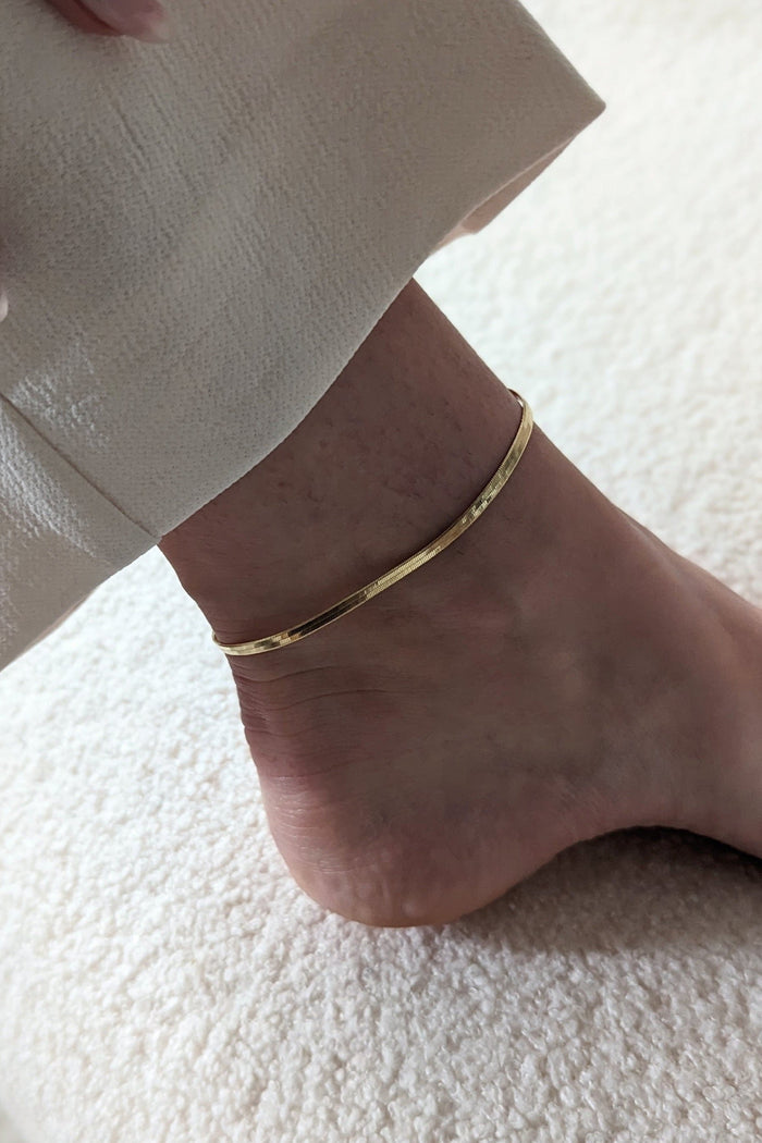 A gold herringbone pattern anklet worn on an ankle, with a beige trouser in the background.