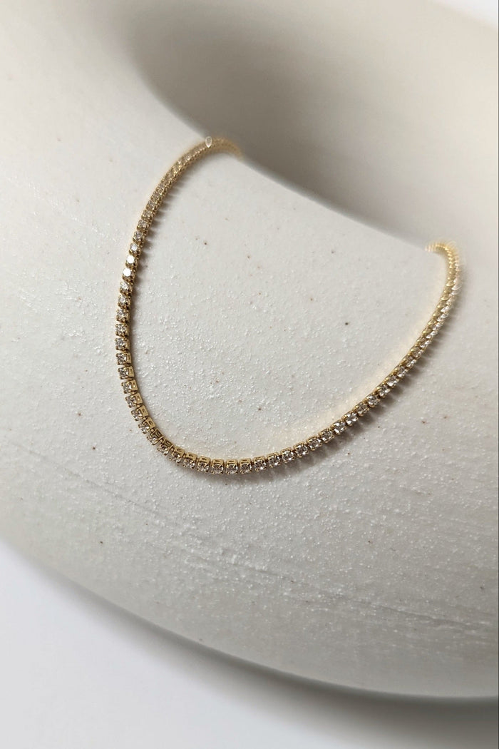 A gold-colored tennis necklace with cubic zirconia stones displayed on a white stand.