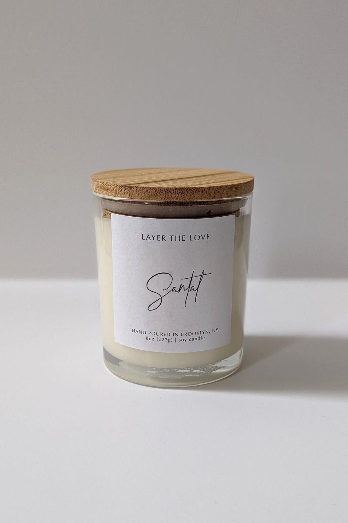 A soy candle in an 8 oz glass jar with a bamboo lid, adorned with hand picked flowers, sitting on a flat surface with dappled light and a shadow of a plant in the background.