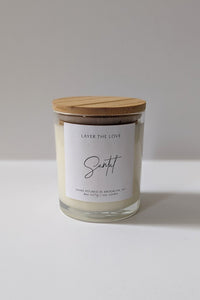 A soy candle in an 8 oz glass jar with a bamboo lid, adorned with hand picked flowers, sitting on a flat surface with dappled light and a shadow of a plant in the background.