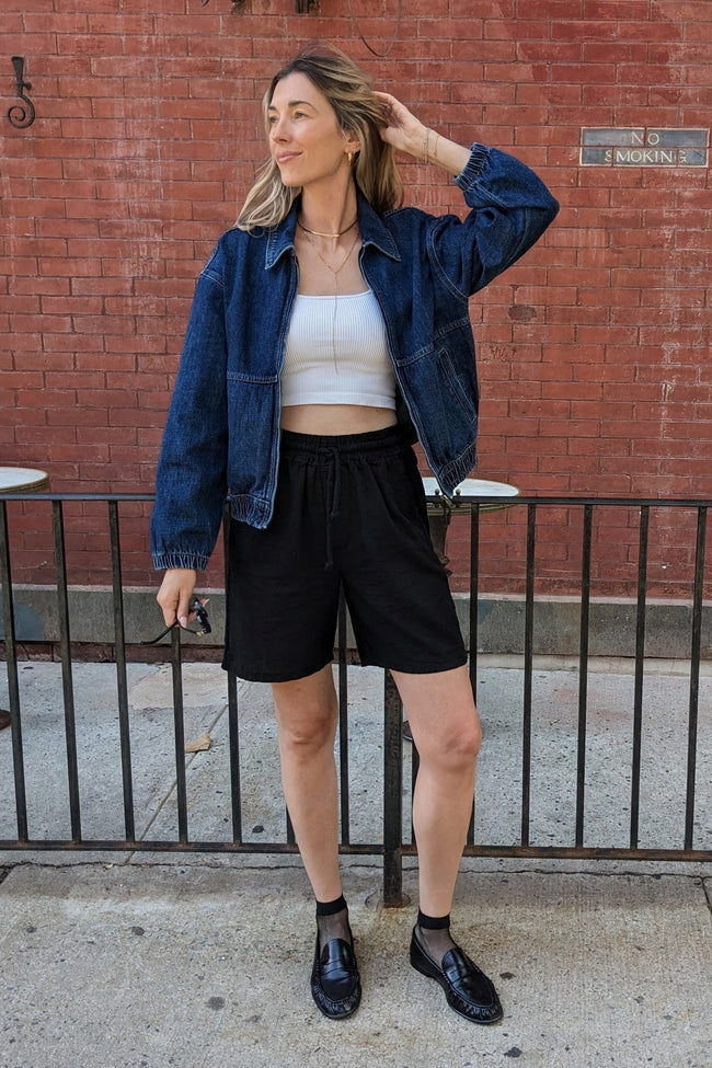 A woman stands on a city sidewalk in front of a red brick wall, wearing a dark blue denim jacket over a white crop top, black shorts, and black loafers.