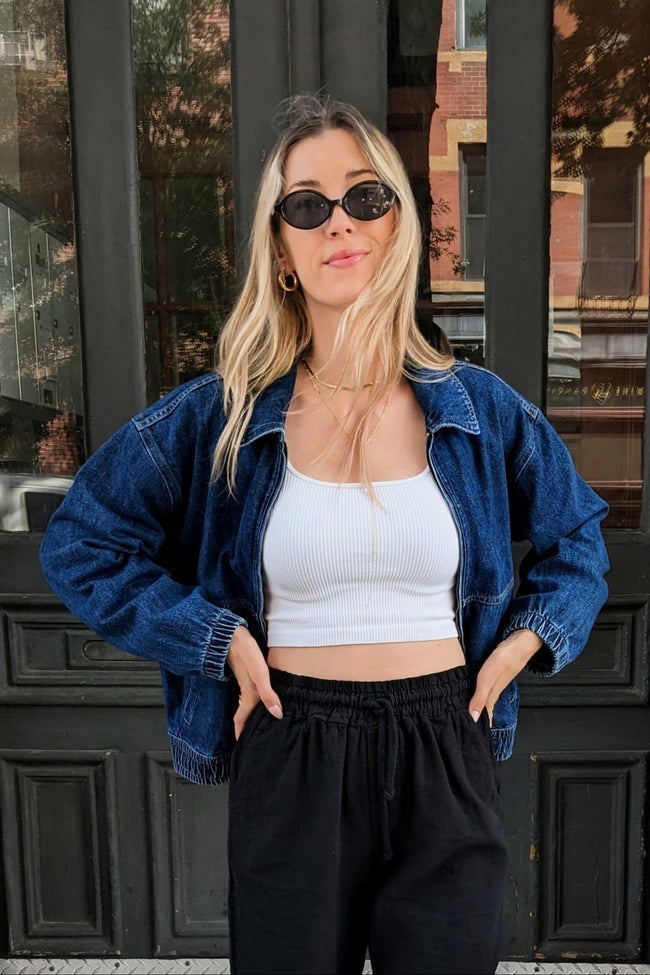 A woman wearing a blue denim jacket over a white ribbed tank top and black sweatpants, standing in front of a dark paneled door.