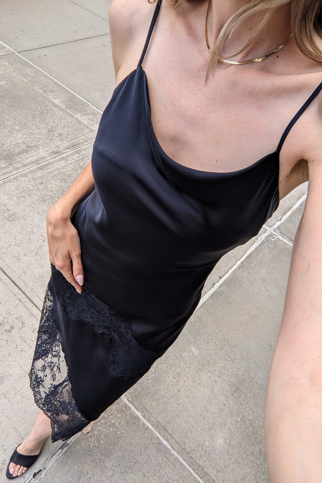 A woman wearing a black satin slip dress with thin spaghetti straps and a black lace hem, photographed in a casual outdoor selfie on a concrete sidewalk.
