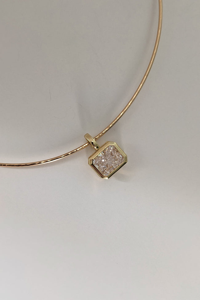 A delicate gold bangle bracelet with a small hexagonal pendant featuring a textured, diamond-like clear stone in a gold bezel setting, displayed on a neutral light gray background.