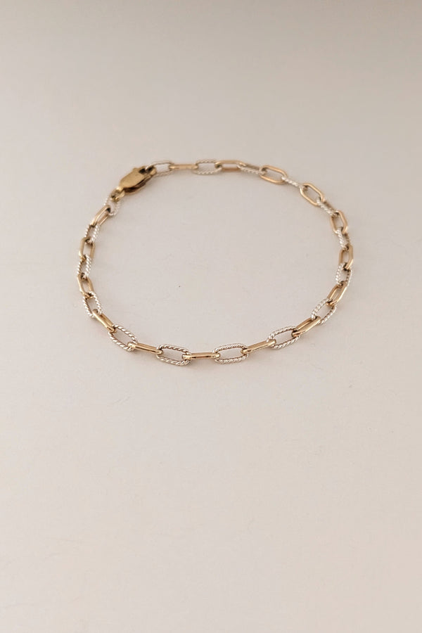A delicate gold chain bracelet with alternating polished and textured oval links, displayed on a plain light beige background.