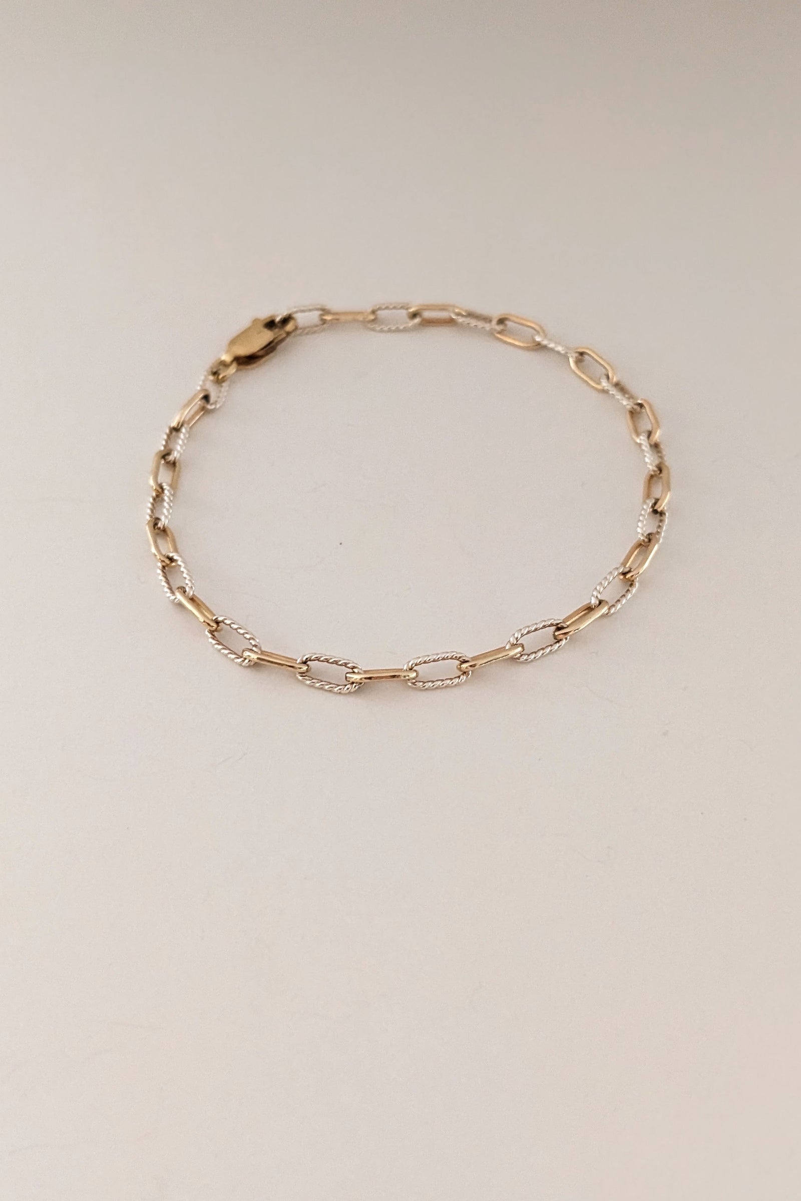 A delicate gold chain bracelet with alternating polished and textured oval links, displayed on a plain light beige background.