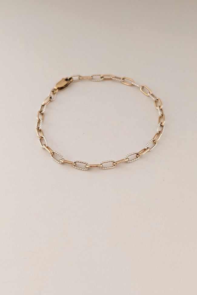 A delicate gold chain bracelet with alternating polished and textured oval links, displayed on a plain light beige background.