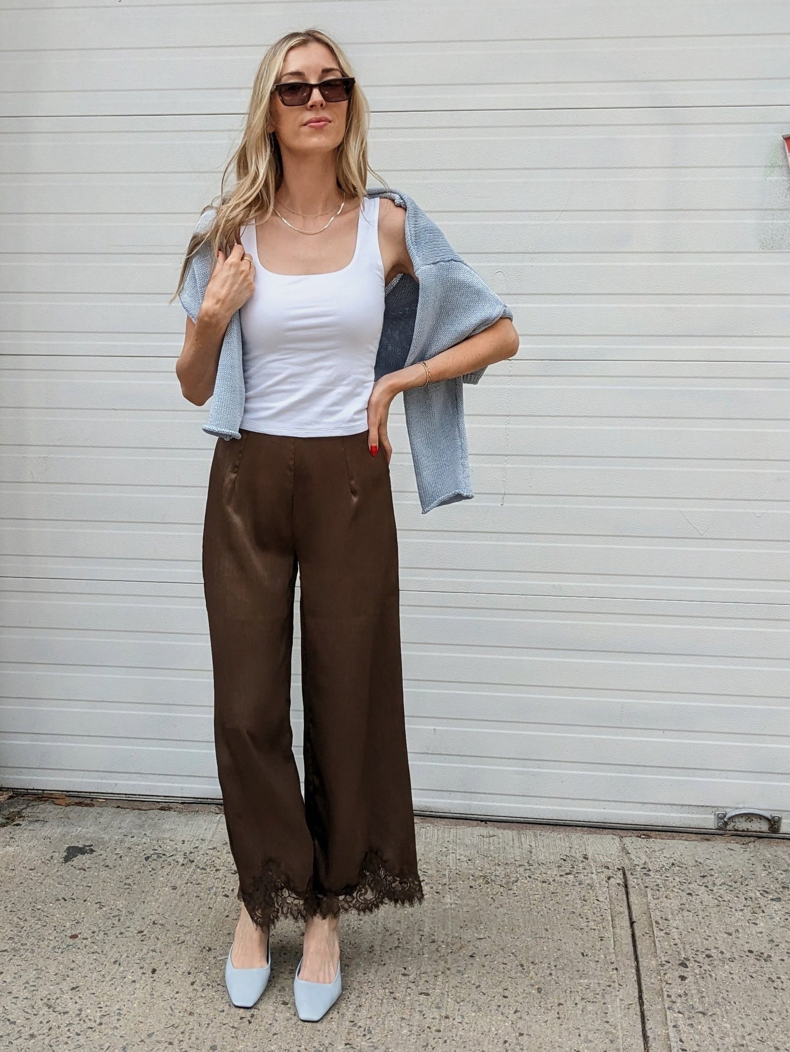 A woman stands outdoors in front of a white garage door wearing a white tank top, brown wide-leg pants with lace trim, a light blue denim jacket draped off her shoulders, and light blue pointed shoes.