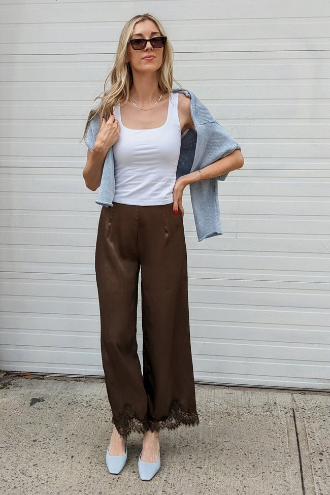 A woman stands outdoors in front of a white garage door wearing a white tank top, brown wide-leg pants with lace trim, a light blue denim jacket draped off her shoulders, and light blue pointed shoes.
