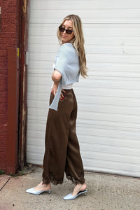 A woman posing outdoors in front of a white garage door and a red brick column, wearing a light blue short-sleeve top, brown wide-leg pants, and white pointed-toe mules.