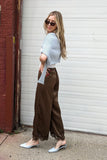 A woman posing outdoors in front of a white garage door and a red brick column, wearing a light blue short-sleeve top, brown wide-leg pants, and white pointed-toe mules.
