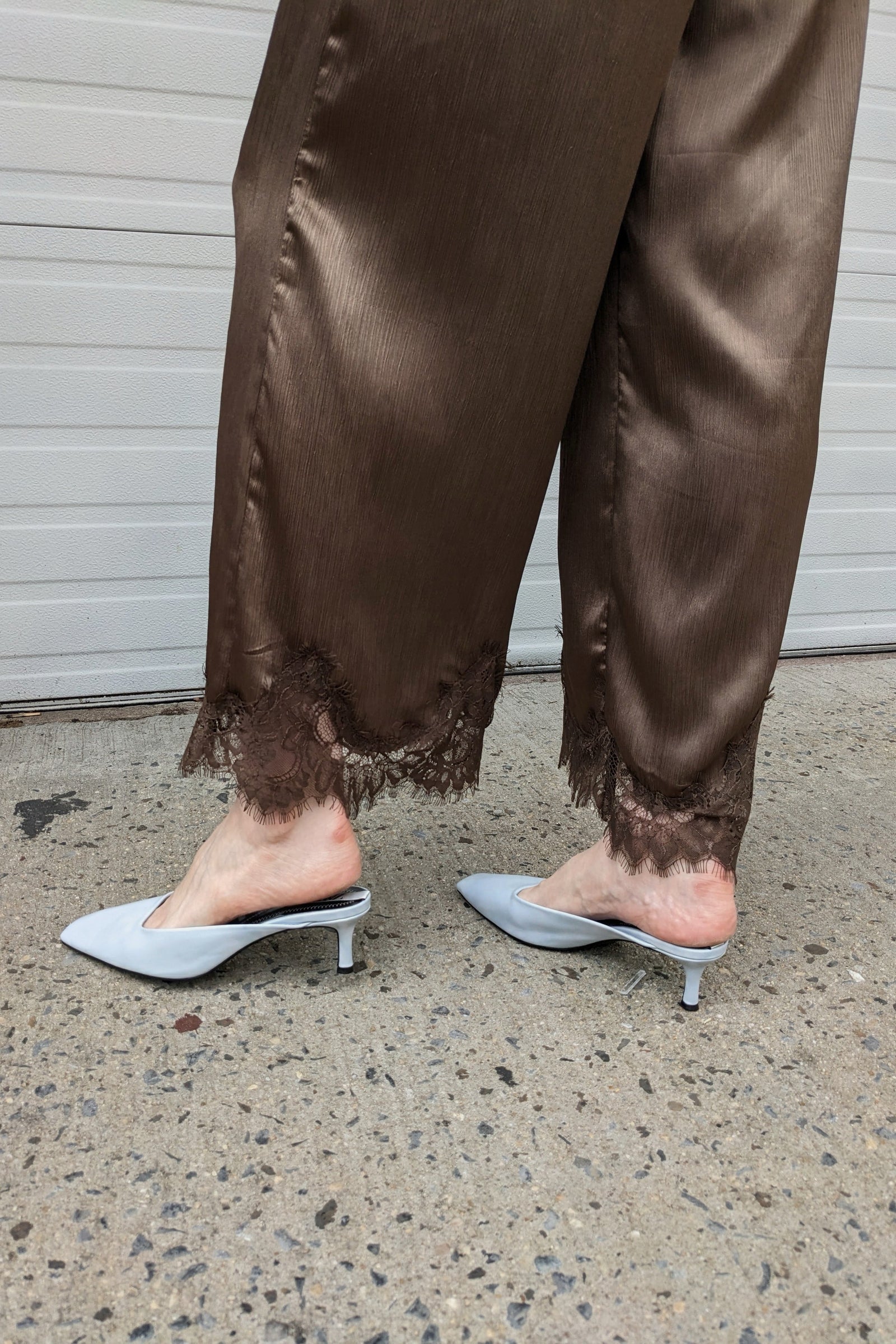 A person wearing brown satin wide-leg pants with lace trim at the hem, paired with light gray pointed-toe mule heels, standing on concrete in front of a white garage door.