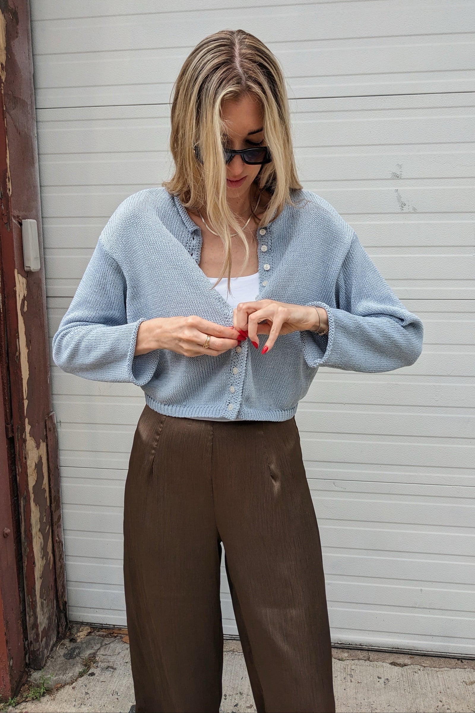 A woman wearing a light blue knit cardigan over a white top and brown wide-leg pants, standing in front of a white garage door.