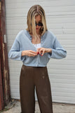 A woman wearing a light blue knit cardigan over a white top and brown wide-leg pants, standing in front of a white garage door.