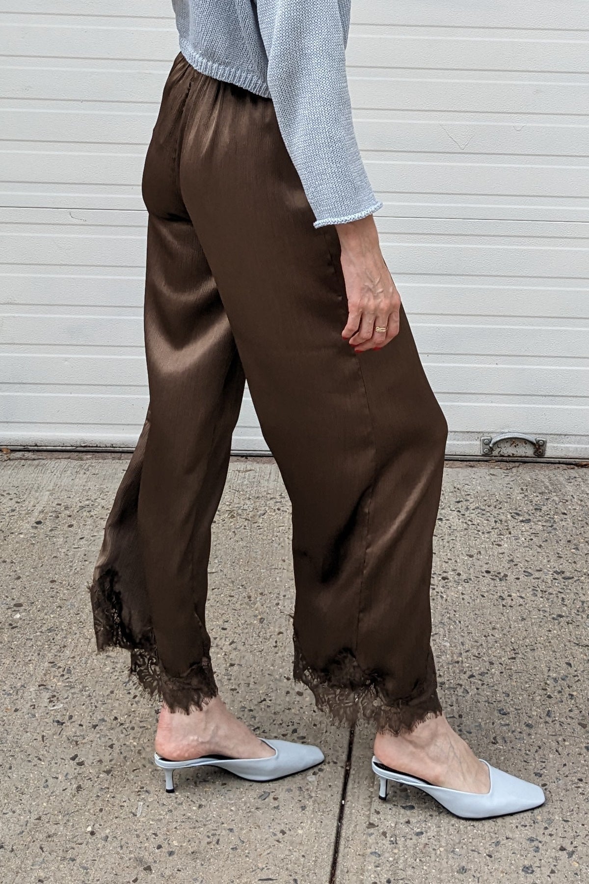 A woman stands in profile on a concrete sidewalk in front of a white garage door, wearing a light blue long-sleeve top, brown satin-like wide-leg pants with a raw hem, and light blue slingback flats.