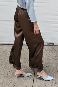 A woman stands in profile on a concrete sidewalk in front of a white garage door, wearing a light blue long-sleeve top, brown satin-like wide-leg pants with a raw hem, and light blue slingback flats.