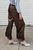 A woman stands in profile on a concrete sidewalk in front of a white garage door, wearing a light blue long-sleeve top, brown satin-like wide-leg pants with a raw hem, and light blue slingback flats.