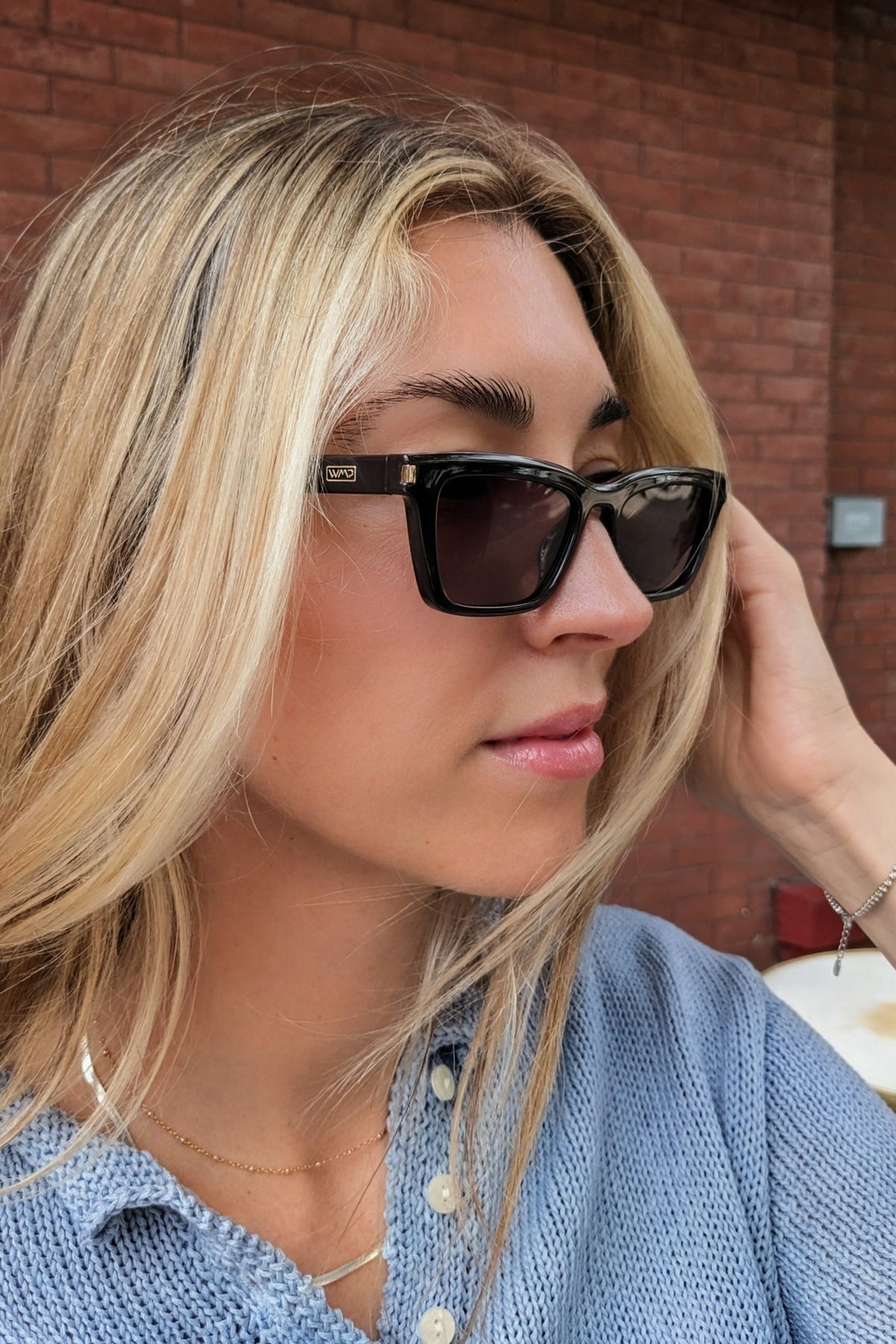 A close-up lifestyle shot of a blonde woman wearing black cat-eye sunglasses with gold hardware and a blue knit top, set against a red brick wall background.