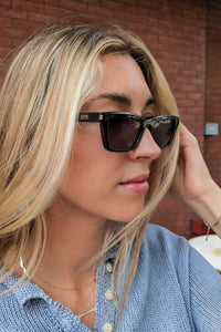 A close-up lifestyle shot of a blonde woman wearing black cat-eye sunglasses with gold hardware and a blue knit top, set against a red brick wall background.