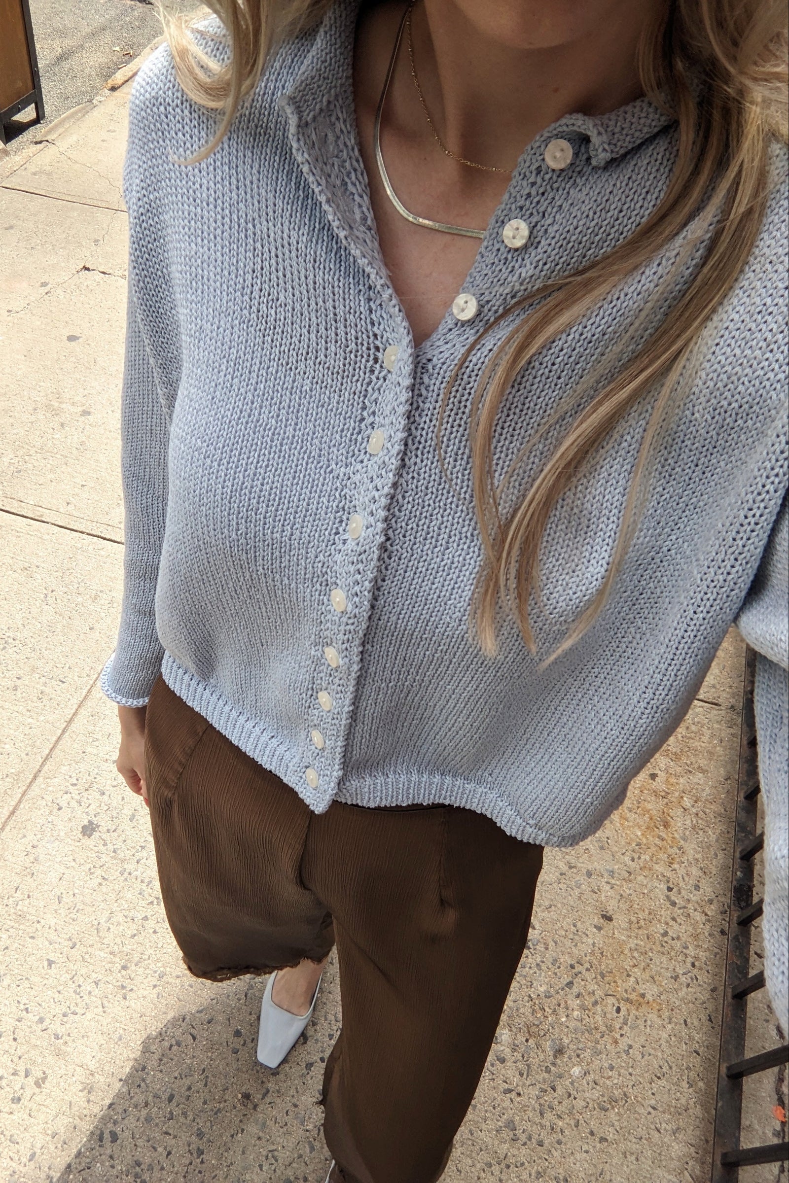 A person wearing a light blue knit button-front cardigan with brown pants, standing outdoors on a concrete sidewalk.