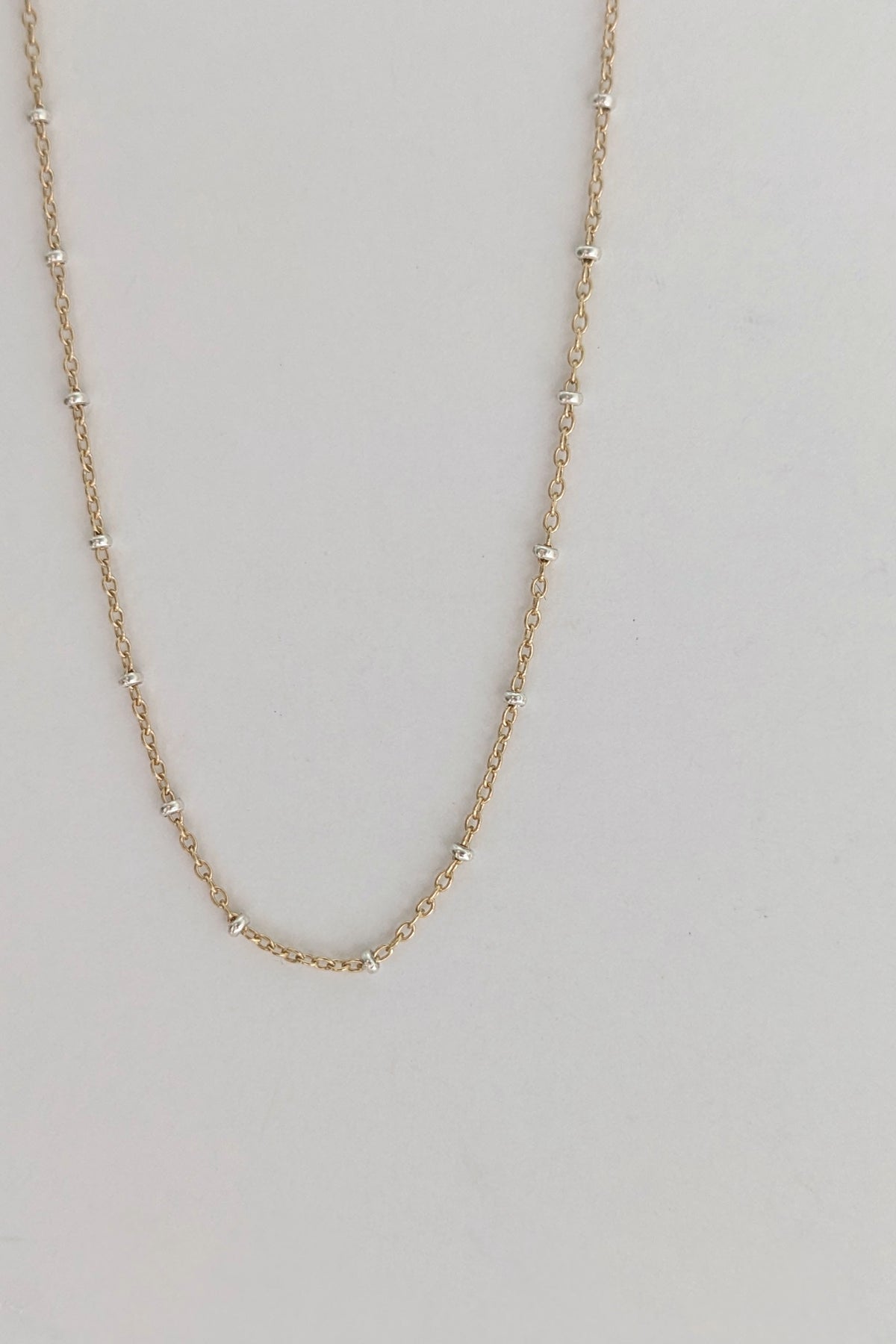 A delicate gold chain necklace with small, evenly spaced silver-toned bead accents, displayed against a plain light gray background.