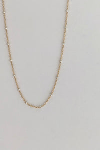 A delicate gold chain necklace with small, evenly spaced silver-toned bead accents, displayed against a plain light gray background.