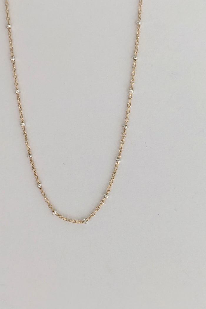 A delicate gold chain necklace with small, evenly spaced silver-toned bead accents, displayed against a plain light gray background.
