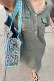A close-up lifestyle shot of a person wearing an olive green linen shirt and carrying a large tote bag with a blue, green, and red botanical pattern and striped handles, standing on a concrete sidewalk.