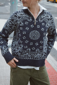 A person wearing a navy quarter-zip sweater with a white paisley and floral bandana pattern, styled with a white collared shirt and olive green pants on an outdoor street setting.